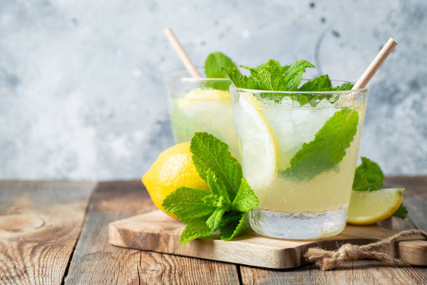 Two glass with lemonade or mojito cocktail with lemon and mint, cold refreshing drink or beverage with ice on rustic blue background. Copy space - Photo, Image