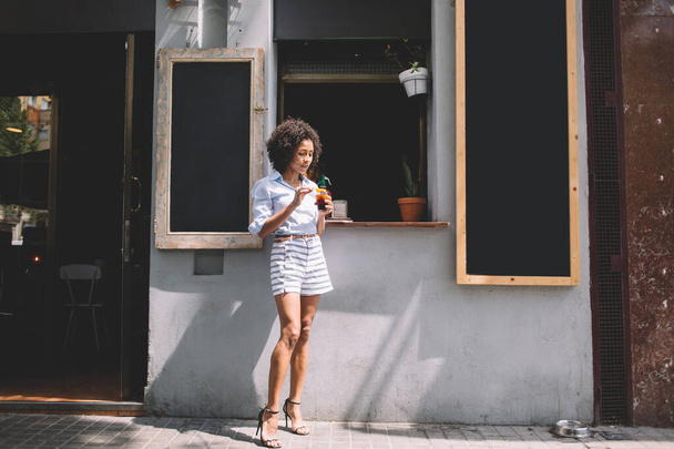Young African American trendy female in shorts and heels with curly hair standing with glass of cocktail outside of street of cafe in sunny day - Фото, зображення