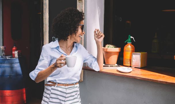 Cheerful young ethnic lady in casual outfit and sunglasses leaning elbow on wooden windowsill and drinking takeaway coffee from cup - Photo, Image