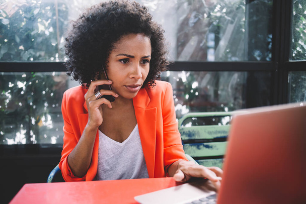Serious African American businesswoman sitting at table and browsing laptop while working with smartphone and listening attentively to colleague on mobile phone - Foto, Bild