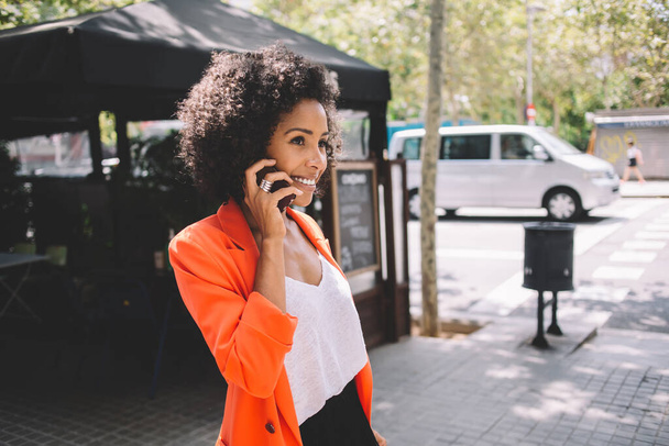 Young African American female in trendy outfit with fluffy hair talking on smartphone while standing on street near small cafe and looking away - Fotografie, Obrázek