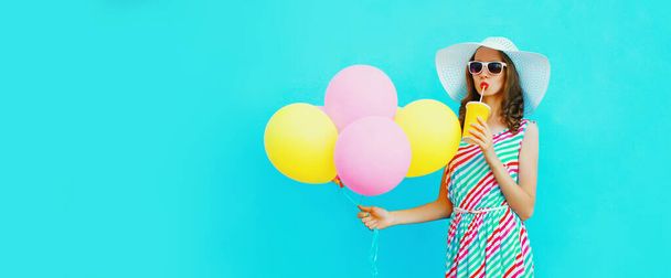 Portrait of beautiful young woman with bunch of balloons drinking a fresh juice wearing a colorful dress on blue background - Foto, Imagen