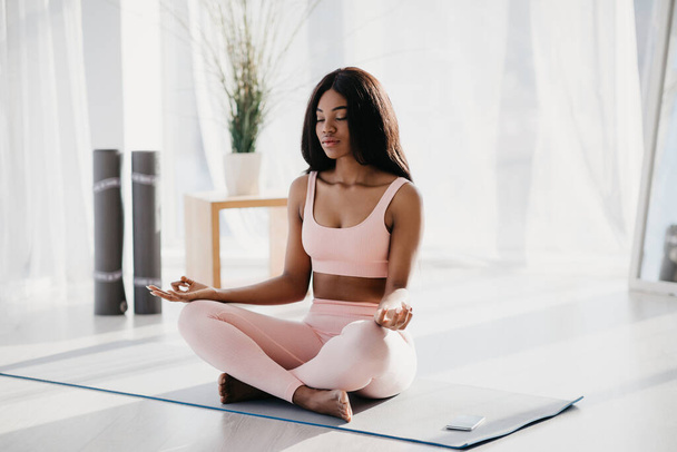 Mujer negra joven y pacífica sentada en pose de loto, meditando, alcanzando la paz interior y la armonía en la alfombra en casa - Foto, Imagen