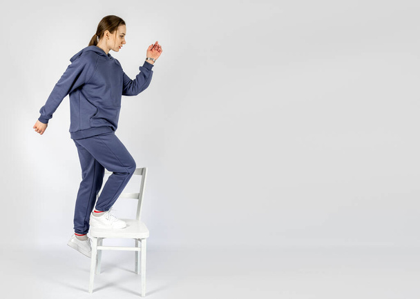 Полнометражный портрет молодой спортивной женщины в синем хлопчатобумажном комбинезоне в толстовке, играющей на белом стуле. Студийный снимок блондинки в капюшоне с длинными волосами. - Фото, изображение