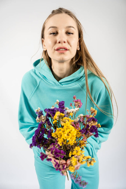 Unaltered candid emotional portrait of young happy blonde long hair woman with flower bouquet on light background. - Фото, изображение