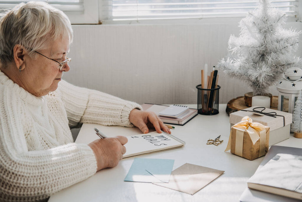 Egészséges újévi fogadalmak idősebb felnőttek számára. Idősebb idős nő fehér pulóverben kézzel írott szöveget ír Céljaimról nyílt jegyzetfüzetben az asztalon. Tervezés és célok kitűzése - Fotó, kép
