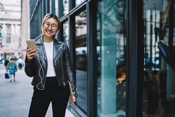 Portrait of joyful Japanese user in earphones and optical glasses using media app for listening radio music on leisure, happy female generation Z with sketchbook and mobile phone posing in city - 写真・画像