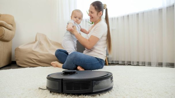 Robot vacuum cleaner helping woman doing chores while she spend time with baby son. Concept of hygiene, household gadgets and robots at modern life. - Photo, Image