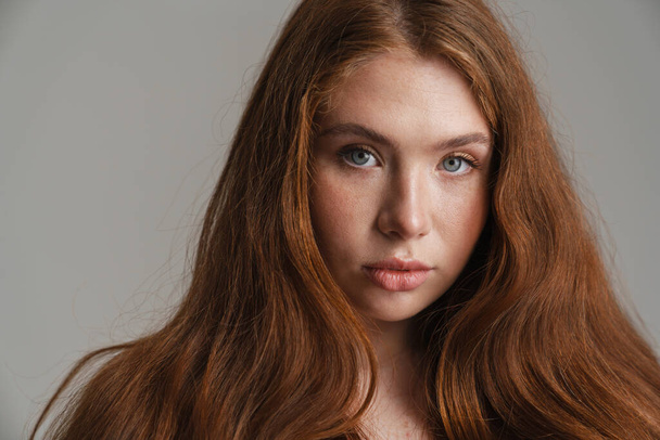 Close up of a young white woman with long red hair looking at camera standing over gray background - Foto, Bild