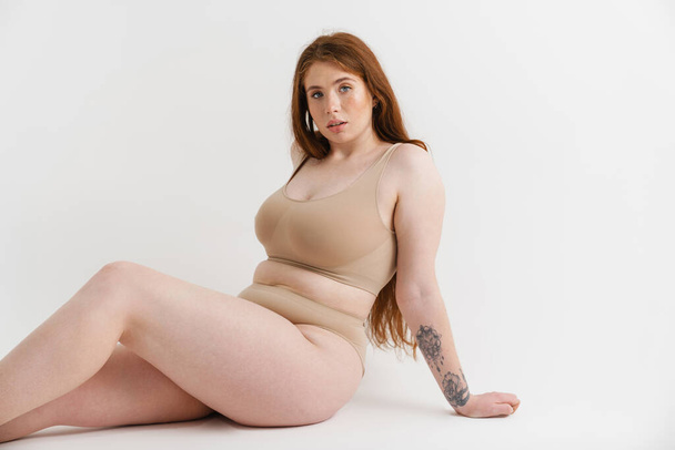 Young white plus size woman wearing lingerie sitting on a floor isolated background posing - Photo, image