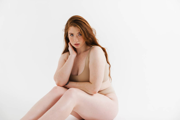 Young white plus size woman wearing lingerie sitting on a floor isolated background posing - 写真・画像