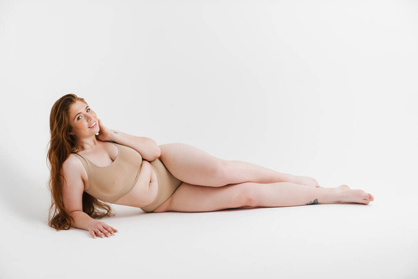 Young white plus size woman in lingerie posing over gray wall background, laying on a floor - Fotó, kép