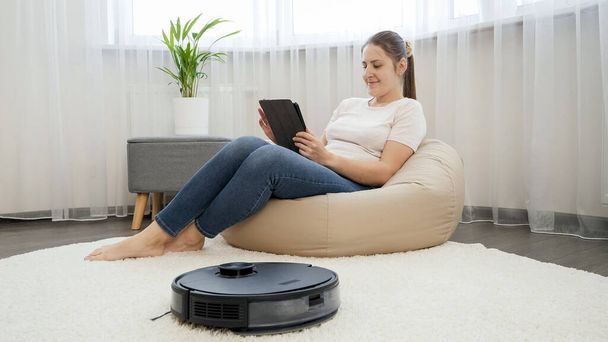 Young freelance woman working in living room while robot vacuum cleaner doing house chores. Concept of hygiene, household gadgets and robots at modern life. - Foto, Imagem