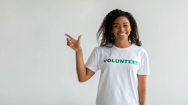 Kijk daar. Gelukkig zwart vrouwelijke vrijwilliger wijzend vinger opzij en glimlachend, tonen vrije ruimte over lichte muur, panorama - Foto, afbeelding