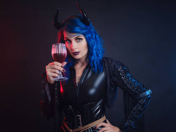 Devil. Funny imp with black horns and latex suit with shibari at Halloween party. Halloween makeup. The devil costume. woman tastes red wine looks like blood in glass - Photo, Image