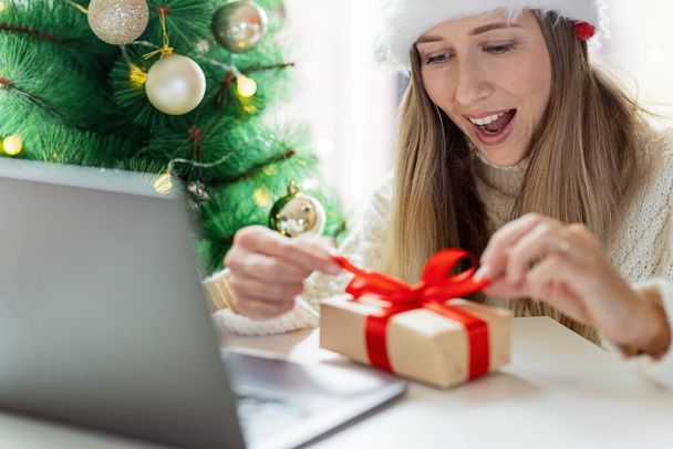 Young woman opening Christmas or new year present when calling to friends via video chat from home - Zdjęcie, obraz