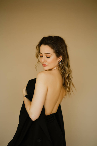 portrait of young beauty caucasian woman with her bare back covered by a jacket , a girl with makeup and curly hair , a dark eyes lady with earrings-rings and bright makeup on light background - Foto, afbeelding