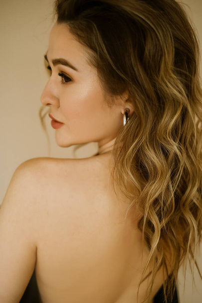 portrait of young beauty caucasian woman with her bare back covered by a jacket , a girl with makeup and curly hair , a dark eyes lady with earrings-rings and bright makeup on light background - Photo, image