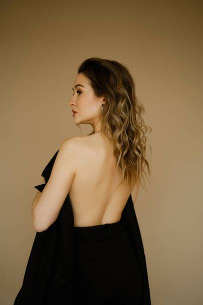 portrait of young beauty caucasian woman with her bare back covered by a jacket , a girl with makeup and curly hair , a dark eyes lady with earrings-rings and bright makeup on light background - Foto, Bild
