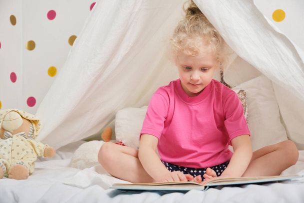 a cute girl with curly hair reads a book in a hut from a sheet on the bed, little girl having fun and playing in her tent, Hut in the kids room - Foto, afbeelding