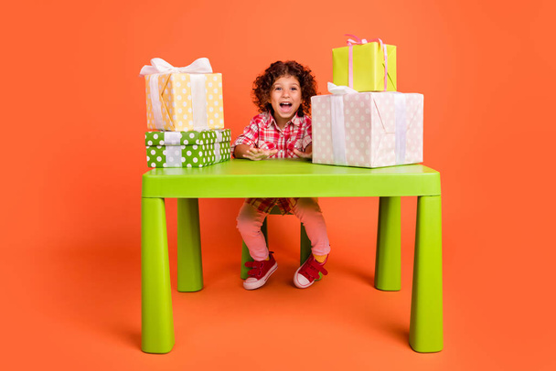 Photo of funny impressed school girl dressed checkered clothes sitting table getting many present isolated orange color background - Foto, immagini