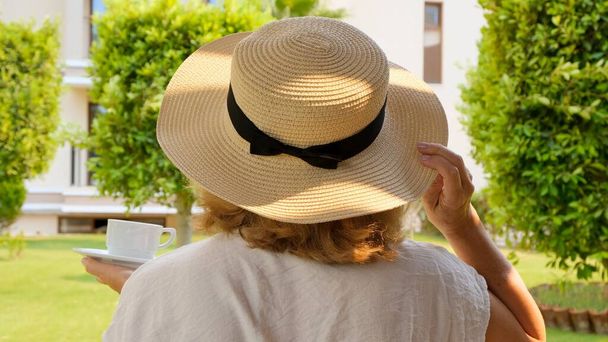 γυναίκα ηλικίας 50-55 ετών σε ένα ψάθινο καπέλο κατέχει ένα φλιτζάνι ζεστό ρόφημα και πίνει καφέ, κάθεται το πρωί σε μια καλοκαιρινή βεράντα σε μια ηλιόλουστη μέρα. Η έννοια της καλημέρα, το ταξίδι και την προσδοκία των νέων - Φωτογραφία, εικόνα