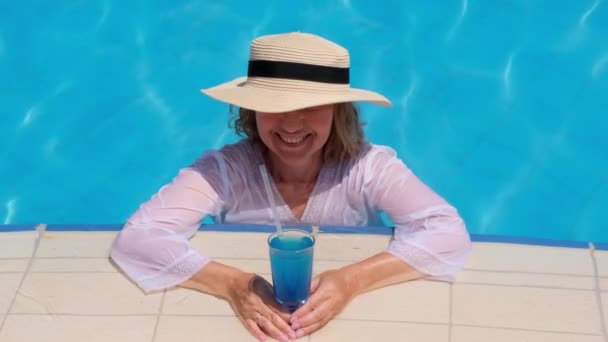 Donna anziana sorridente che si rilassa vicino alla piscina blu all'aperto con un cocktail blu. La gente si sta godendo le vacanze estive - Filmati, video