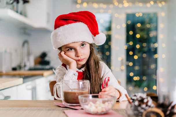 Bored cute little girl kid in Santa Claus hat sitting on the decorated for winter holidays modern kitchen with cocoa drink. Festive season. Copy space. - Foto, afbeelding