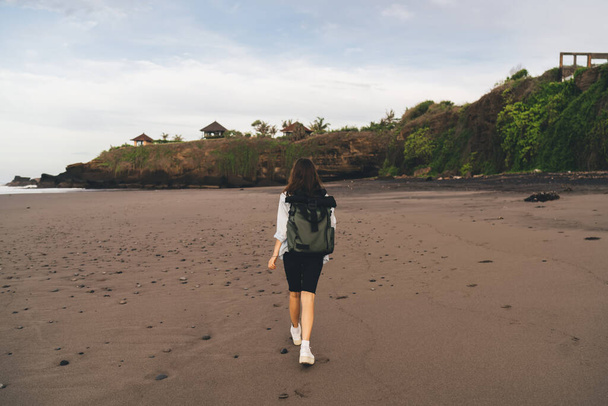Achteraanzicht van het hele lichaam anonieme vrouwelijke toerist in casual kleding met rugzak wandelen op zandkust in de buurt van golvende zee - Foto, afbeelding