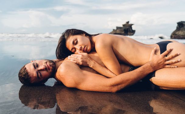 Side view of man and ethnic woman in black swimwear laying on sandy beach near waving sea and embracing each other - Photo, Image