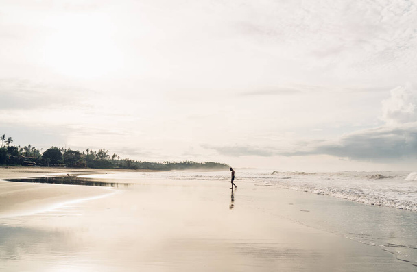 Side view of unrecognizable man walking on wet sandy shore near waving sea under bright shining sun and enjoying harmony - Foto, Bild