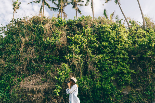 Low angle of side view of young woman in hat standing near high green hill and taking photo on photo camera - 写真・画像