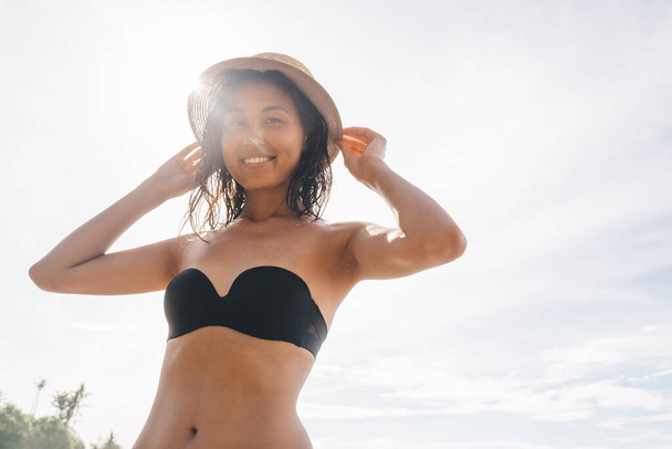 Low angle of cheerful ethnic female in bikini and hat smiling and looking at camera while standing on sandy beach against cloudless sky - Zdjęcie, obraz