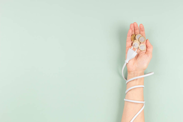El cable de alimentación eléctrica envuelve la mano de la mujer para alcanzar monedas de dinero en euros. Eficiencia energética, consumo de energía, aumento del precio de la electricidad y concepto de energía caro - Foto, imagen
