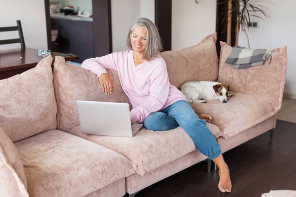Older mature woman using wireless laptop apps browsing internet sit on sofa, smiling middle aged female working distantly on computer surfing web communicating online looking at screen at home - Foto, afbeelding