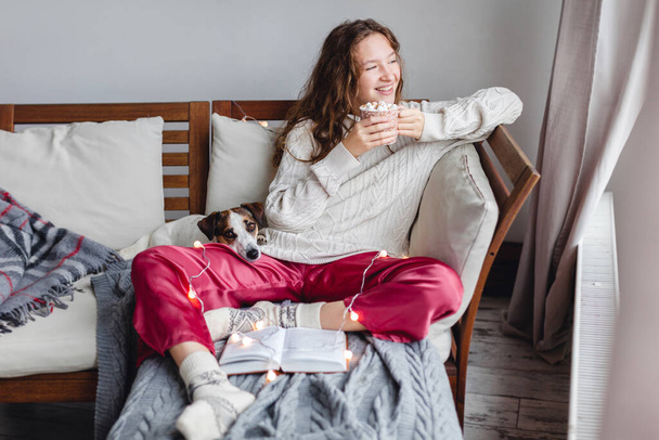 Cold autumn or winter weekend Happy woman with dog drinking warm cocoa or coffee with marshmellows. Lazy day in knitted socks on the couch. Cosy scene, cozy home concept - Foto, Imagen