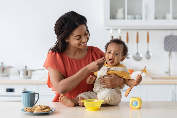 Cuidado de niños. Loving negro mamá alimentación su bebé hijo desde cuchara en cocina - Foto, Imagen