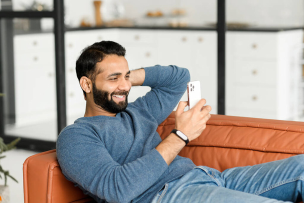 Hilarious 30s Indian man using smartphone sitting on the couch at home - Foto, Imagen