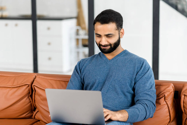 Uomo etnico ottimista in abbigliamento casual utilizzando il computer portatile seduto sul divano - Foto, immagini