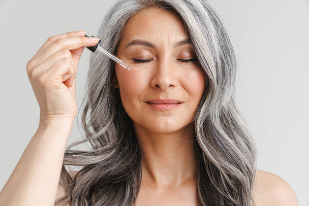 Mature shirtless woman with grey hair applying face serum isolated over white background - Foto, Imagen