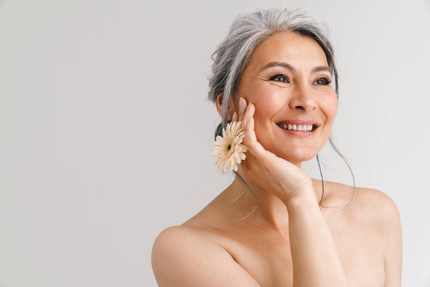 Shirtless woman with grey hair smiling while posing with gerbera isolated over white background - Photo, image
