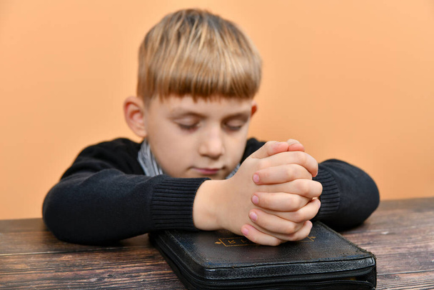 Хлопчик молиться на колінах, склавши руки на закритій Біблії. - Фото, зображення