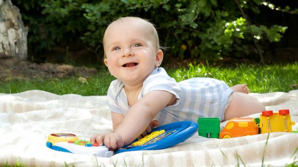 庭で草の上に寝そべってカラフルなおもちゃで遊んでいるかわいい笑顔の赤ちゃん。子供の早期開発、教育、屋外でのリラックスの概念. - 写真・画像
