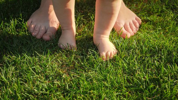 母と新鮮な緑の芝生の上に立つ裸足の赤ちゃんの閉鎖。健康的なライフスタイル、子供の発達と子育ての概念. - 写真・画像