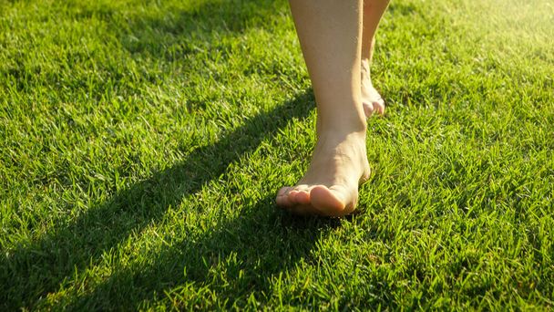 Крупный план взрослых ног, идущих по свежей зеленой траве на фоне заката - Фото, изображение