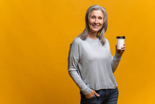 Smiling senior woman holding paper cup of coffee isolated over yellow background - Фото, изображение