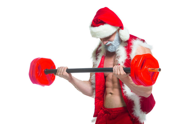 Christmas. Strong muscular Santa Claus with grey beard wearing christmas hat and red shorts is lifting a heavy barbell in a gym performing a workout isolated on white background. - Photo, Image