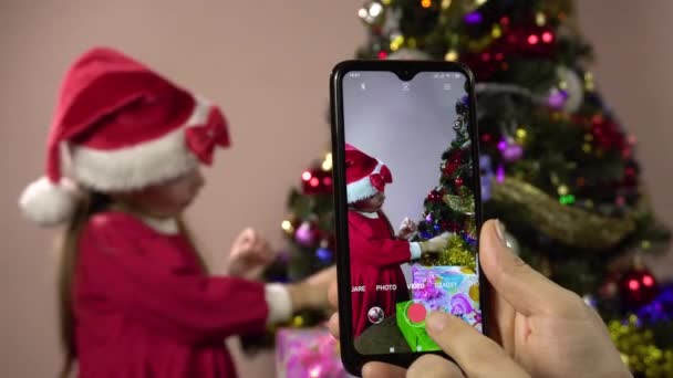 Ein kleines Mädchen als Weihnachtsmann verkleidet schmückt einen Weihnachtsbaum und posiert für Shootings auf einem Smartphone. - Filmmaterial, Video