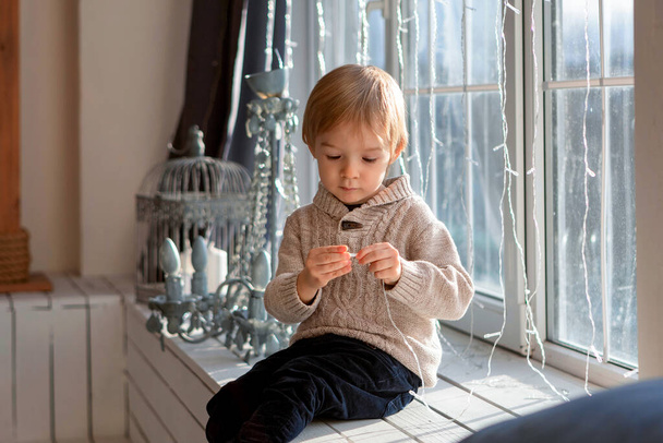 A blond-haired boy is sitting near the window - Фото, изображение