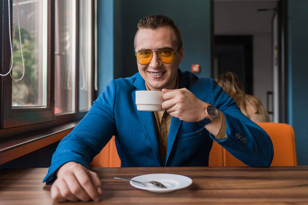 Un hombre de negocios sonriente positivo un retrato elegante de la apariencia caucásica en gafas de sol, chaqueta y camisa, se sienta en una mesa en una pausa de café en un fondo de café. - Foto, Imagen